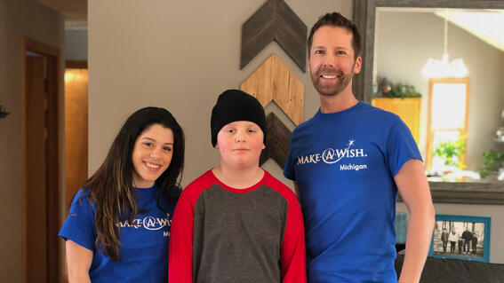 Two adults in royal blue Make-A-Wish t-shirts and jeans stand on either side of a child in a red and gray shirt and black beanie. The background is a living room with a gray couch and wooden wall art.