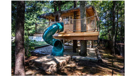Libby's wooden treehouse nestled among the trees, with a thatched roof and a green twisty slide