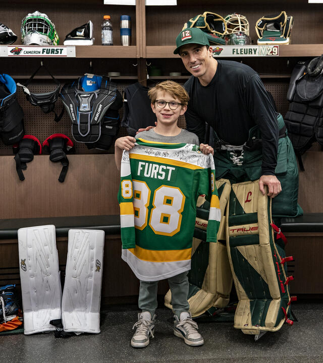 Cameron holding up a custom jersey next to Marc Fleury in the Wild locker room