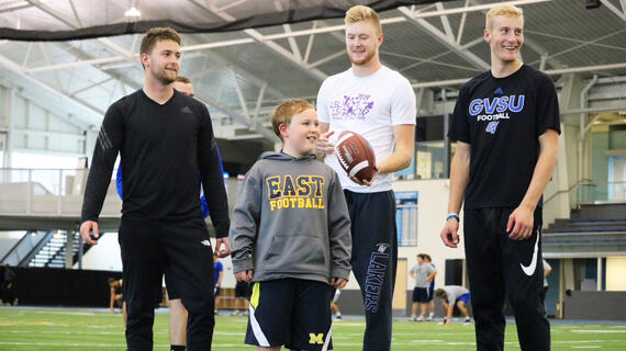 Three college football players pose for a photo on the football field with a young blond child wearing a gray hoodie and black shorts. The player in the middle is holding a football. 