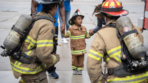 Little Jayce dressed in his fire fighter outfit standing with other fire fighters