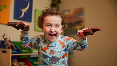 Dressed in gray pajamas covered with multi-colored dinosaurs, a 4-year-old child pretends to be a dinosaur roaring. In the background is a playroom, with a shelf full of toys and dinosaur images on the walls. 