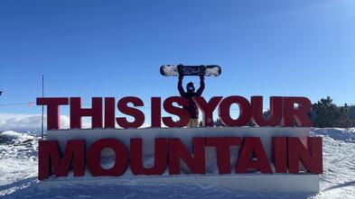 girl on snowy mountaintop holding snowboard above her head while standing behind a sign that reads, THIS IS YOUR MOUNTAIN
