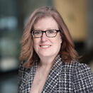 Woman with  reddish blond hair and glasses, wearing grey checked blazer