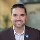 A man smiling. He has dark brown hair, a mustache, and a beard. He is wearing a brown sports jacket with a Make-A-Wish star pin.