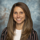 Kelly LaVergne headshot, woman smiling wearing red and blue striped blazer on neutral background 