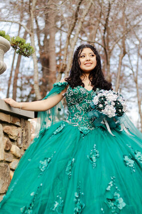 Emely standing outside with her hand extended onto a tall stone pillar wearing a green ballgown