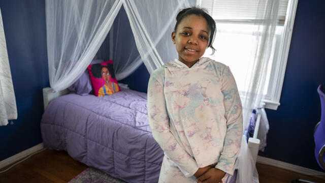 A child wearing a pastel tie-dyed hoodie with a unicorn design and matching pants stands with hands clasped in front of a bed decorated with sheet white curtains and a purple bedspread.