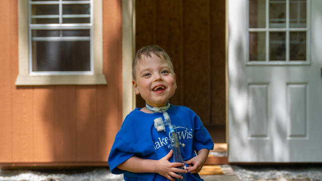Axle is standing in front of his new playhouse. He is smiling while holding his breathing tube.